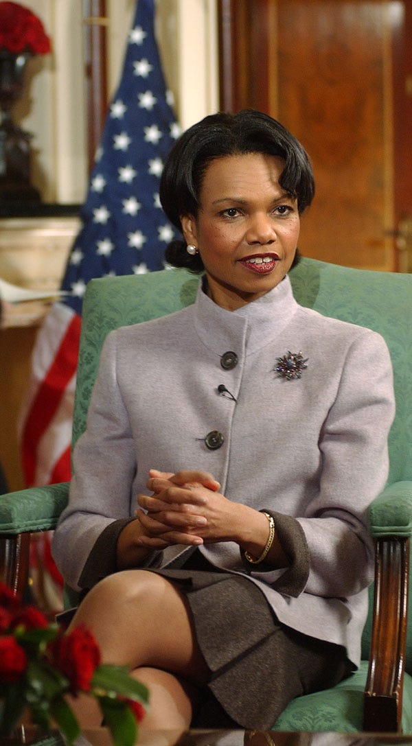 A woman with brown skin and black hair sits in a green armchair in front of an American flag. She wears a gray jacket and speaks with her hands folded.