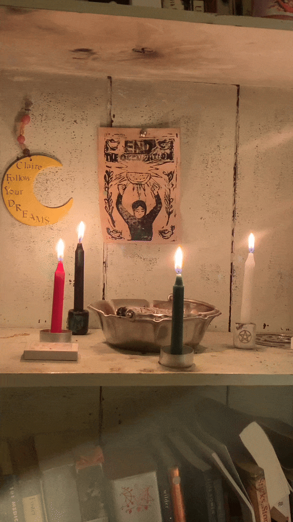 4 ritual candles — red, black, white and green stand aflame around a metal burn bowl on a bookshelf. Above the burning candles, an ink-blot print reads “End the Occupation” with the figure of a person holding a watermelon slice.