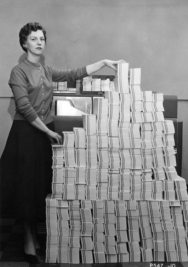 A woman standing by a huge stack of punch cards — almost as tall as she is!