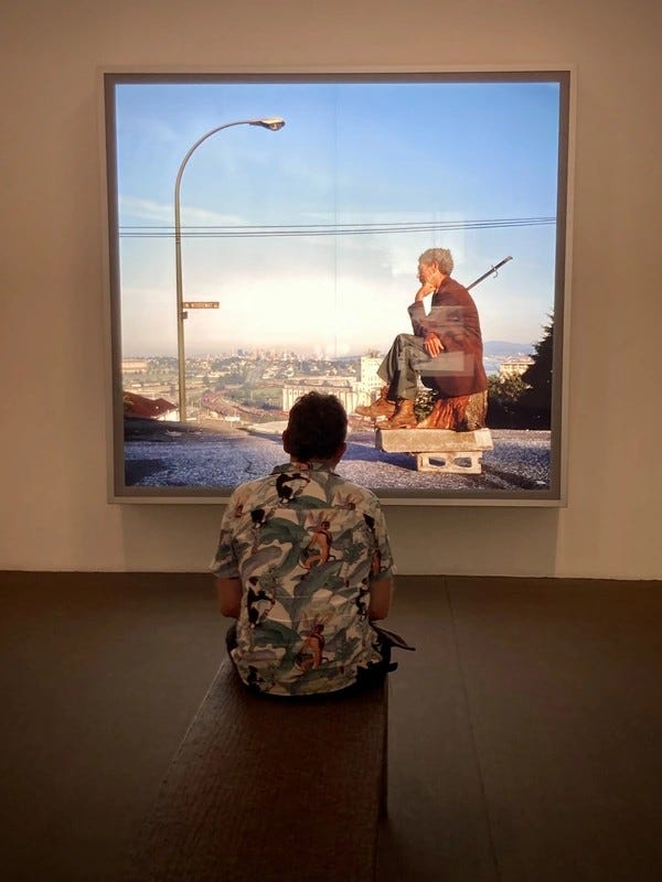 À La Virreina Centre de la Imatge à Barcelone, une personne vêtue d’une chemise à fleurs est assise sur un banc en bois devant une grande photographie encadrée de Jeff Wall. L’œuvre s’intitule The Thinker (1986) et représente un homme sur fond de paysage urbain dans la position bien connue du chef-d’œuvre de Rodin… #SilentSunday © 20240811 Gilles Denizot