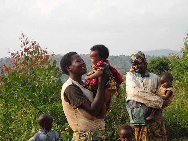 A woman stands while lifting a baby up to her face. She is smiling at the baby. There is another woman in the background holding a young child on her hip, looking at the woman in the middle of the image and smiling. They are all standing outside in among some green bushes, with hills in the distance.
