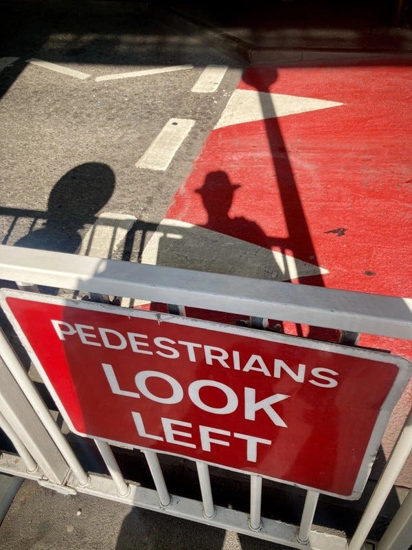 #Off2Europe À LHR, un panneau rouge et blanc indique « PEDESTRIANS LOOK LEFT » (piétons, regardez à gauche) avec une rambarde métallique et les ombres d’une personne en arrière-plan… #SilentSunday © 20240818 Gilles Denizot