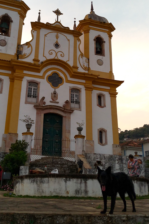 Em primeiro plano, um cão preto de porte grande olha para a câmera. Em segundo plano, é possível ver a mureta de uma fonte que está desligada e uma senhora mexendo em uma mangueira com as mãos. Em terceiro plano, a fachada de uma enorme igreja aparece.