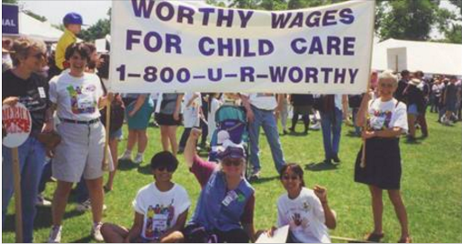 People gathered around holding a banner, "Worthy Wages For Child Care 1-800-U-R-Worthy"