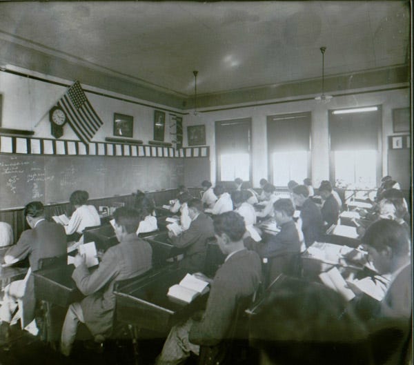 A black and white photo depicts high school aged students of Indian descent sitting in rows of desks in a school classroom. There is a United States flag placed above the chalkboard at the front of the room.
