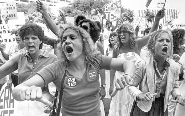 Several women hold signs, march, and shout. The woman in the foreground wears an “ERA YES” button on her shirt.