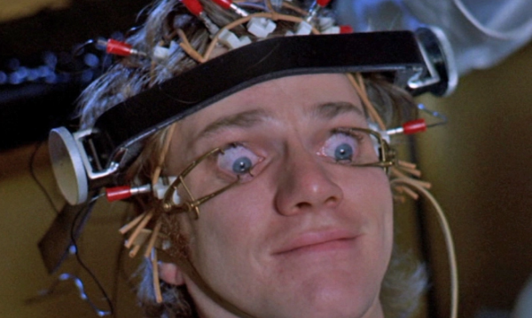 Screengrab from the movie “A Clockwork Orange” by Stanley Kubrick. In the screen there is the face of a white young man with electrodes on his head and with his eyes forced to be opened. He looks distressed.