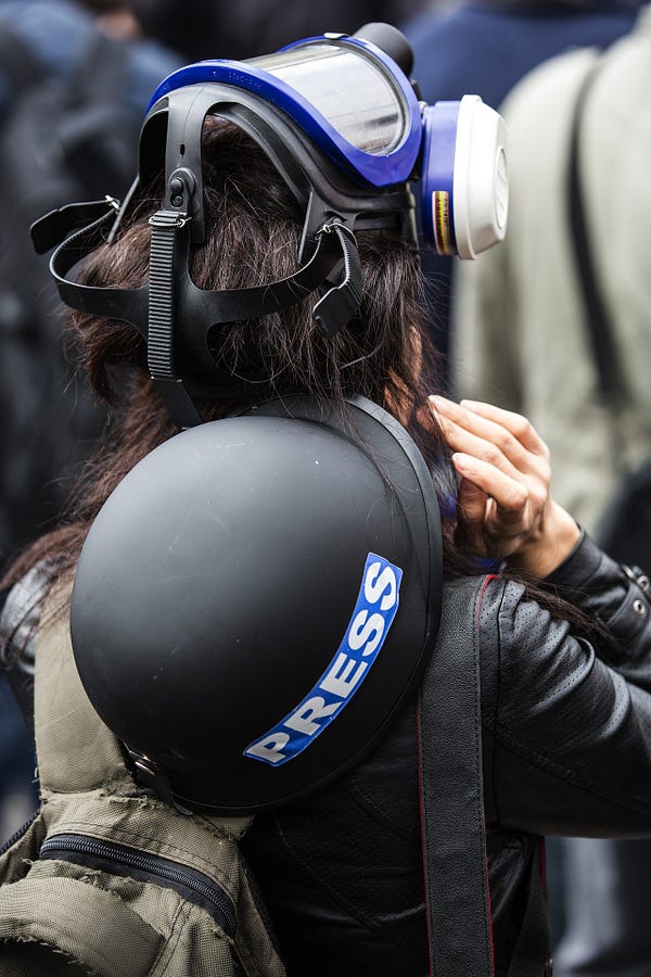 Woman with long hair, a tear gas mask and a shrapnel-proof helmet with a blue and white “PRESS” sticker on it from behind.