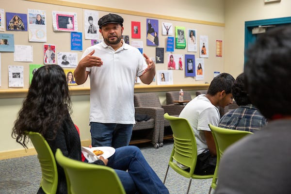 Michael Lozano stands and talks to a woman sitting in a green chair at a library room. Teens artwork are displayed in the background and a few other teens are sitting in the room.