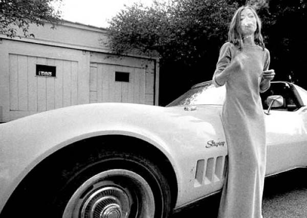 A black and white photo of young Joan Didion smoke a cigarette while leaning on a 1970’s Stingray Corvette.