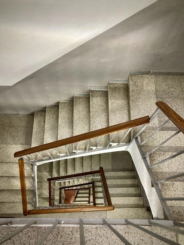 Vue en contrebas d’un escalier en colimaçon aux marches grises et à la main courante en bois, formant un motif géométrique vertigineux #SilentSunday Perspective © 20240804 Gilles Denizot