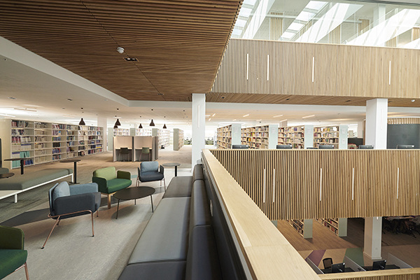Various furniture and tables on the left with bookcases in rows in the far back. On the right, a spacious area for stairs where you can look up or down to other floors.