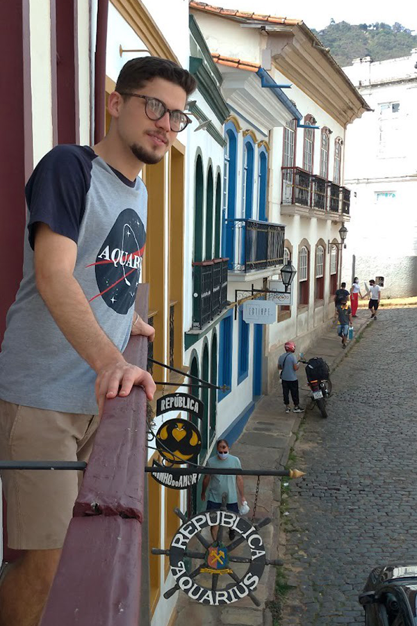 Homem branco está à esquerda da foto, apoiando as mãos em uma sacada no segundo andar de uma casa de estilo colonial barroco. É possível ver outras casa semelhantes em segundo plano, assim como pessoas andando na rua abaixo da sacada. Uma placa redonda que se assemelha a uma roda de leme diz: “República Aquarius”, com um brasão no meio.