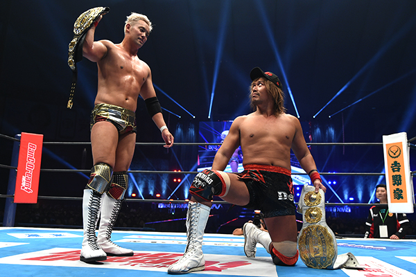 Kazuchika Okada stands above a kneeling Tetsuya Naito in the middle of a ring in an arena. Both have athletic builds and are wearing wrestling gear. They glare at each other as each clutches their title belt.