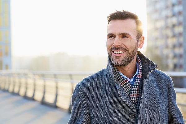 A middle-aged man dressed in a wool coat and scarf stands outside in the city smiling