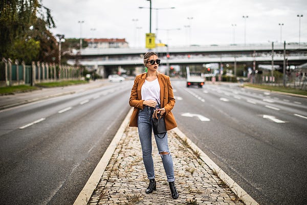 A young lady standing in the middle of the road with a brown jacket and skinny jean on.