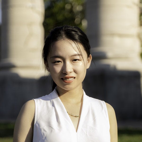 Portrait of AfroLA’s first community engagement fellow, Heather Wang. She’s smiling at the camera in a white top with stone columns in the background.