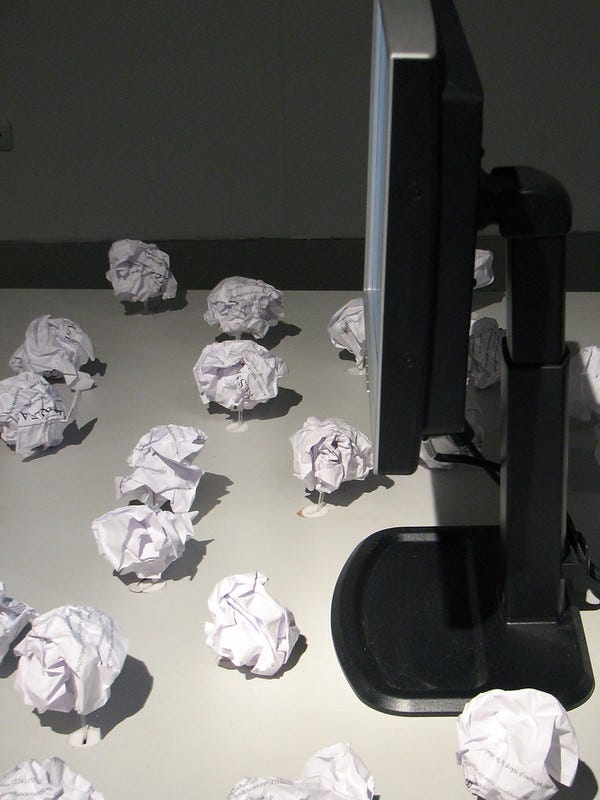 A black computer monitor facing left on a white tabletop with about a dozen balls of crumpled paper scattered in front of it.