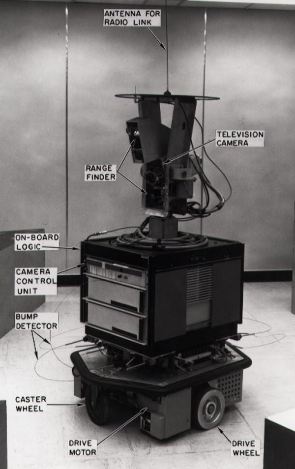 An old school robot made up of old computer parts, circa 1960s. It is labelled with ‘drive wheel’ pointing at wheels, ‘drive motor’, ‘bump detector’, ‘camera control unit’, ‘on-board logic’