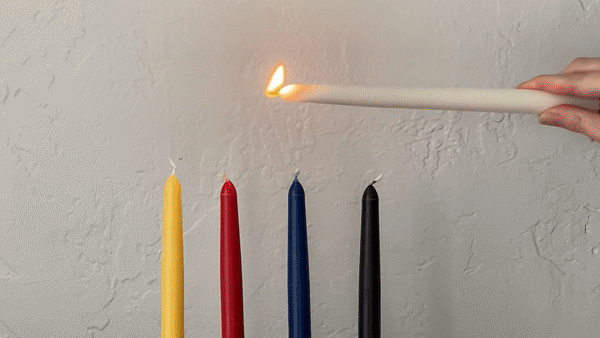Person lighting yellow, red, blue, and black candlesticks with a white candlestick.