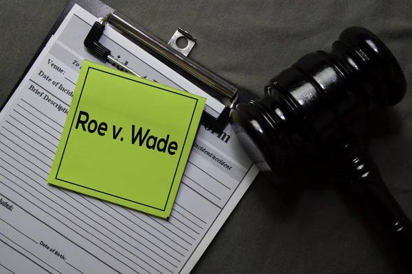 A medical clipboard with a blank form for a medical patient with a green sticky note with bold black caption of Roe v. Wade. Next to clipboard is a dark gavel. All of these items are lying against a gray background