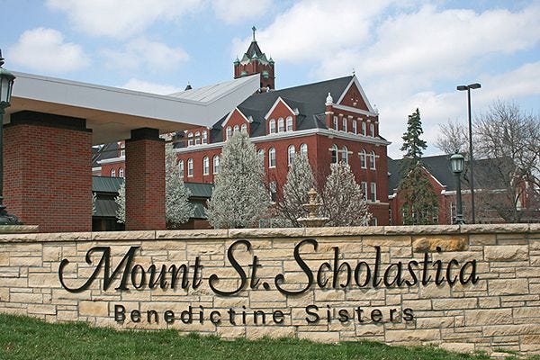 A view of the red brick monastery Mount St. Scholastica in Atchison, Ks surrounded by a short stone wall that says Mount St. Scholastica, Benedictine Sisters.