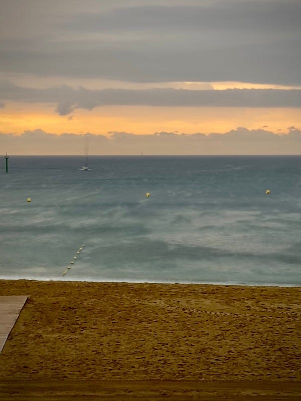 Une scène de plage sereine à Barcelone avec un ciel nuageux, des vagues calmes, un voilier et des bouées jaunes flottant au loin.