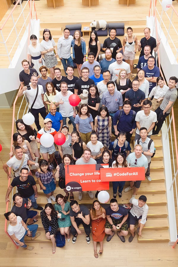 Group of people standing on stairs, smiling at the camera with balloons and signs saying "Change your life" "Learn to code" and "#Codeforall"