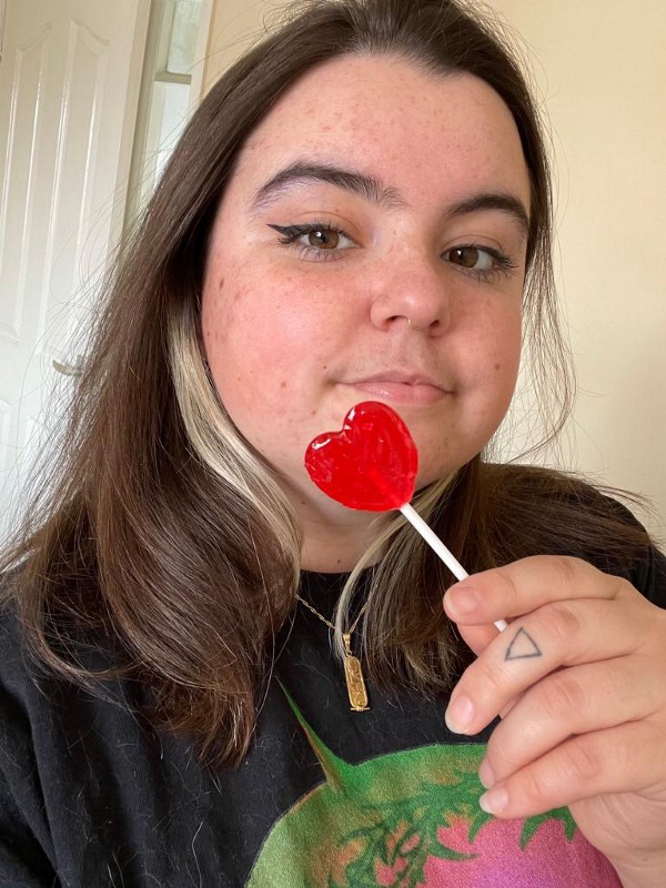 Pip, a white non-binary person with long brown hair and brown eyes, holds up a red heart-shaped lollipop while looking at the camera.
