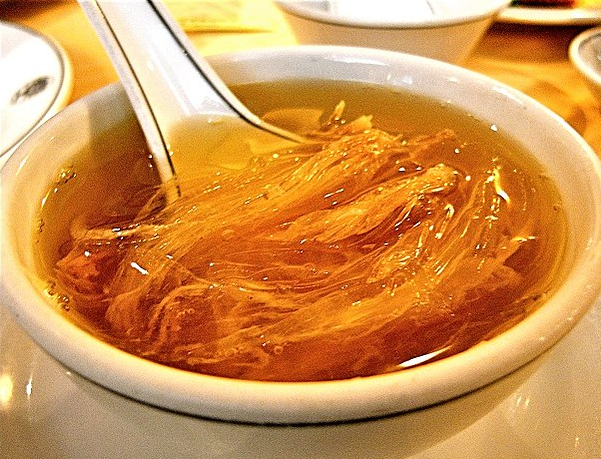 A picture of a Shark fin soup in a bowl. The broth is clear or slightly amber, with a silky and smooth consistency. Floating in the soup are thin, translucent strips of shark fin, which have a gelatinous texture similar to soft noodles or strands of jelly. These strips may be interspersed with shreds of chicken, pieces of crab meat, or other ingredients like mushrooms or bamboo shoots, adding more texture and variety to the dish.