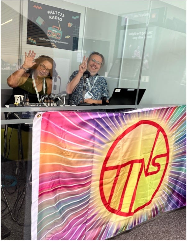 Photograph of woman and man waving at camera from behind glass as they give a radio show. In front of the glass is a large colourful flag with the letters ‘TTNS’ in a large circle in the centre.