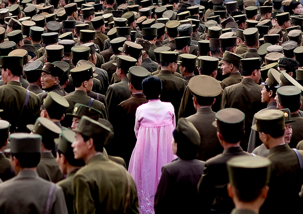 A woman stands in a crowd of North Korean soldiers wearing a pink Hanbok