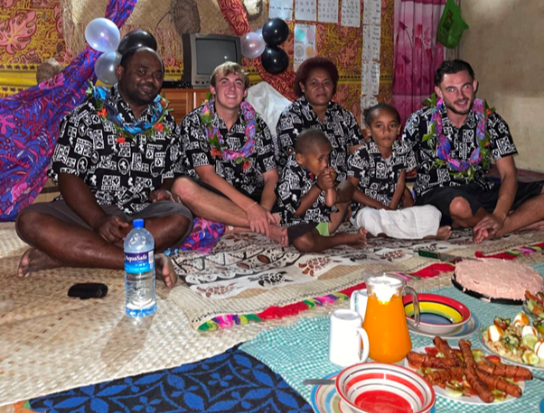 Corey sits with his Fijian family and another Laidlaw scholar. They all are wearing matching traditional Fijian shirts and floral garlands.