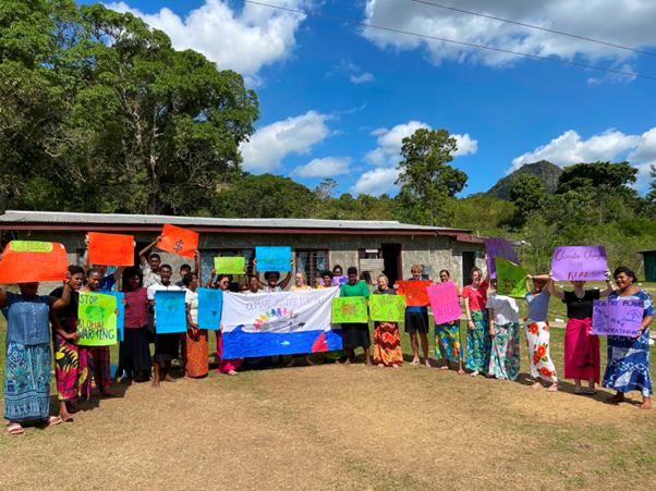 A big group of Laidlaw scholars and their Fijian hosts stand smiling and holding brightly coloured placards. Behind them is a smal building, large green trees and a bright blue sky.