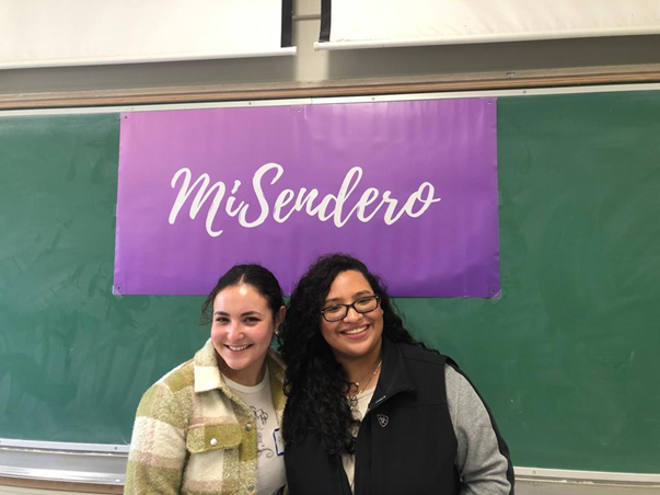 Two young women, both with olive-brown skin tones, smiling in front of a purple MiSendero flag in front of a green classroom board. The girl on the right has long curly hair and is wearing glasses. The girl on the left has her hair tied back in a ponytail and is wearing a checkered chartruste top.