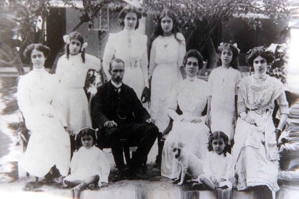 A black-and-white photograph shows a man in Victorian looking clothes, surrounded by many women in Victorian dresses, all seated for a family portrait.