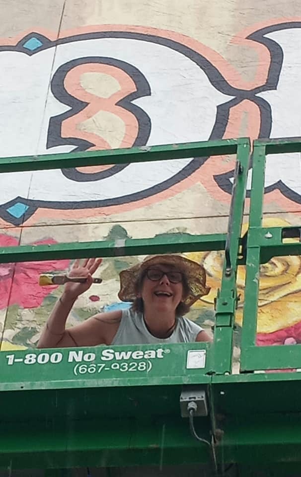 One of the mural artists, while on the lift, smiles and waves to the camera with a brush in hand!