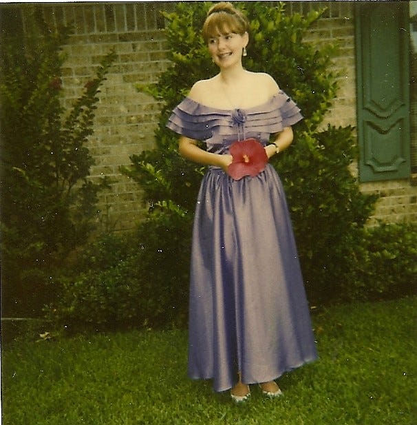 A redhead girl with her hair up wears a long, lavender, off-the-shoulder dress and holds a large red hibiscus flower in front of a huge bush in front of a ranch-style house.