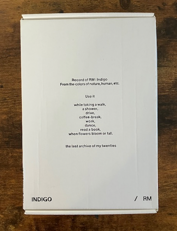 Image shows a physical copy of RM’s 2022 album, Indigo. It is a white box with black writing. The text on the box reads: Record of RM: Indigo From the colors of nature, human, etc. Use it while taking a walk, a shower, drive, coffee-break, work, dance, read a book, when flowers bloom or fall. the last archive of my twenties. INDIGO / RM