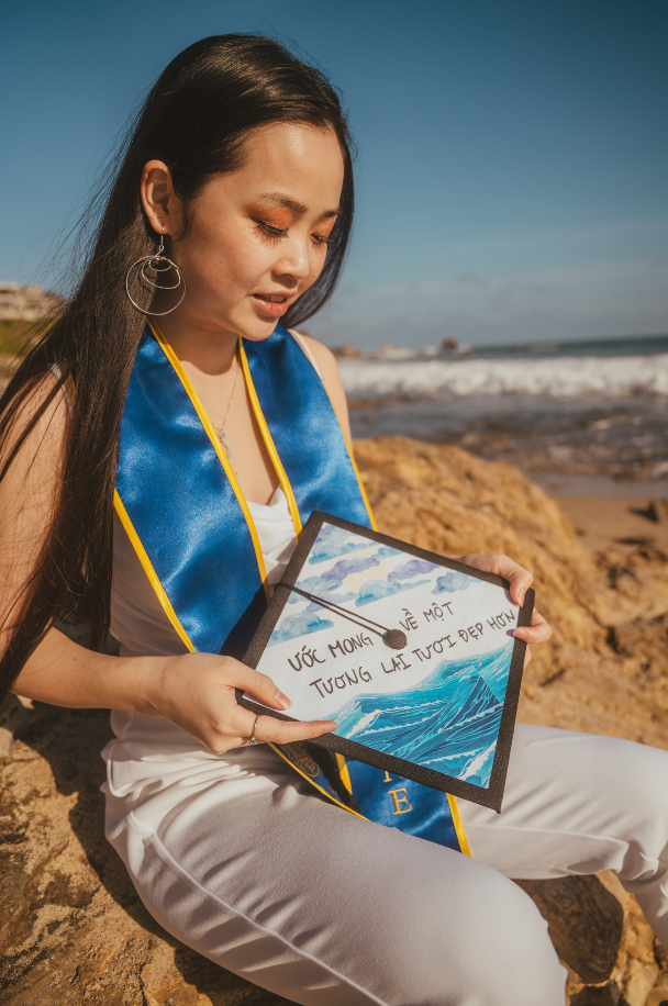 A Vietnamese American woman wears a sleeveless white jumpsuit with a bright blue, gold-lined stole from her university. She is sitting on a big rock at the beach with a decorated grad cap held in her lap. The grad cap has waves on the bottom and clouds with the sun peeking through on the top. In the middle reads “Ước mong về một tương lai tươi đẹp hơn” in Vietnamese, which means “Hoping for a brighter future.”