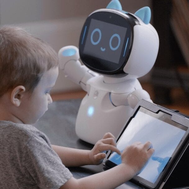 A child typing on a computer tablet sitting next to a foot tall, friendly looking robot with a digital smiling face.