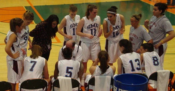 A photo of women on a basketball court, huddled around their couch during a timeout.