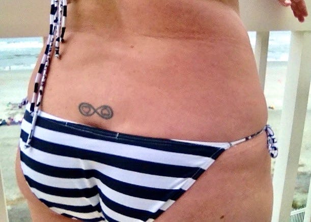 Color photo of the author’s lower back and dark-blue with white striped string-bikini-clad buttocks, while showing her tattoo just above the middle of her bum-crack: a black ‘infinity’ with 2 black-outlined red hearts, one facing up and the other facing down.