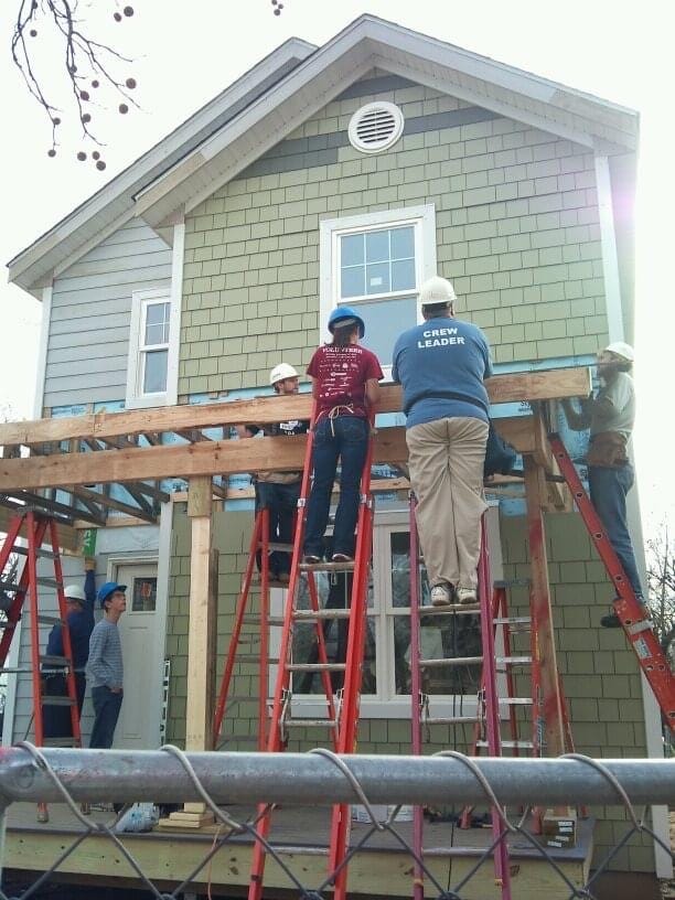 People with construction helmets stand on ladders, constructing a porch roof in front of a nearly completed Habitat for Humanity house. One is the author’s younger daughter. (Photo by author.)