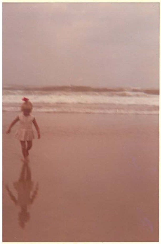 A young female child walking on the beach with her reflection on the water in front of her and the waves in the background