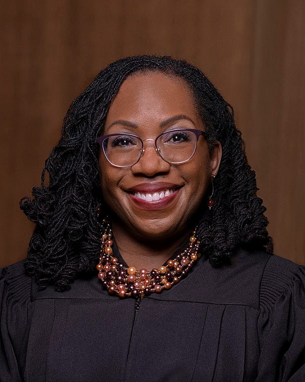 Justice Ketanji Brown Jackson looks at the camera and smiles. She wears a black robe, glasses, and a beaded necklace.