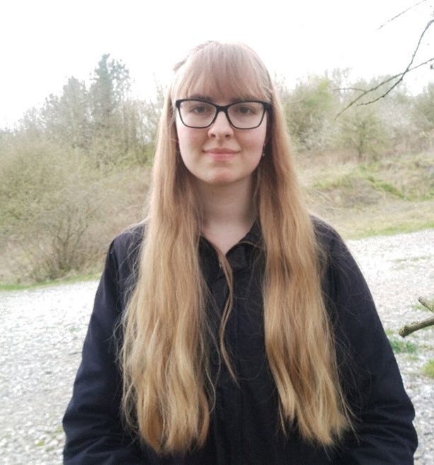 Photo of caucasian woman with long blonder hair, glasses and wearing a black top in front of a background of trees and a river.