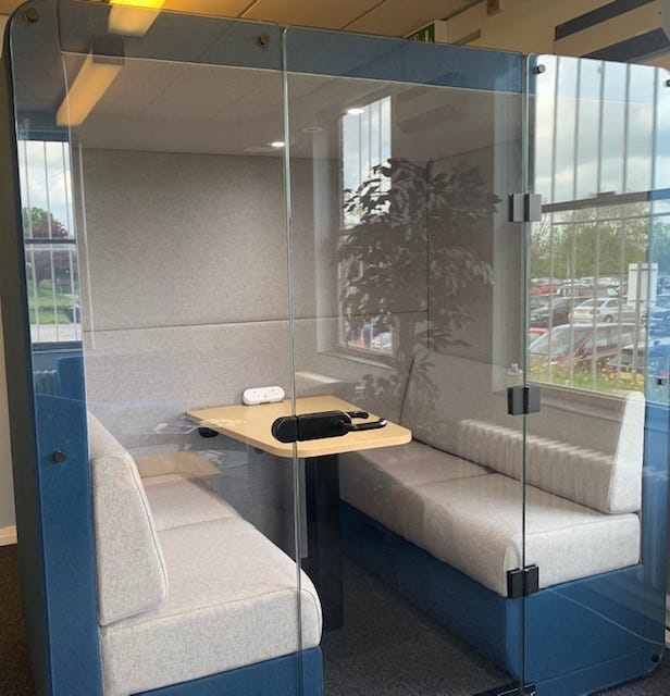 Image of the interior of the quiet pod in the library at Bolton NHS Foundation Trust, showing small table and seating area; a tall green artificial plant is reflected in the glass.