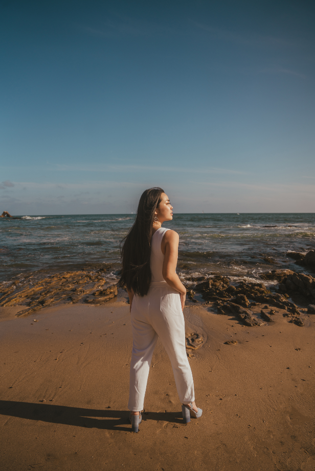 A picture of a Vietnamese American woman standing with her back to the camera, in front of a beach with calm waves. The picture is warm toned. She’s wearing a white jumpsuit with baby blue platform heels, and looks off into the sunset to her right.