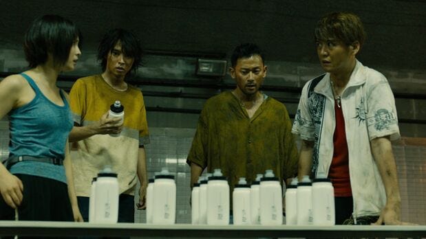 A woman and three Japanese men stand in front of a table with lines of water bottles.